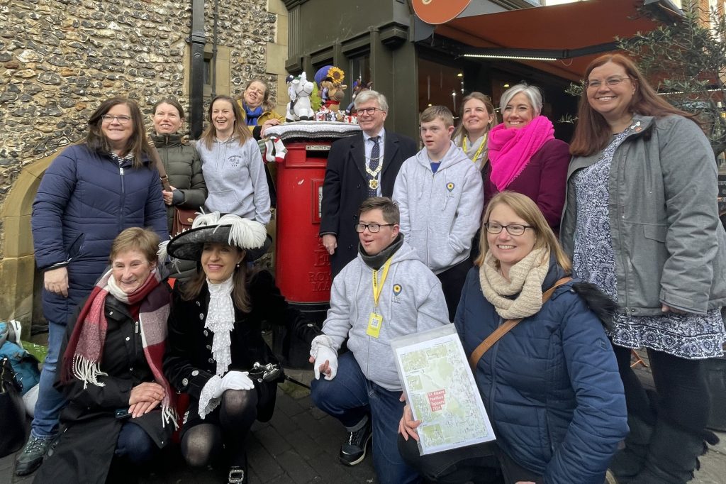 The launch of the 2025 Postbox Toppers fundraiser in St Albans with Clare at the front with her map, and the town centre Postbox at the back, in the middle. It is surrounded by the charity groups, topper makers, dignitaries and Daisy Cooper MP.