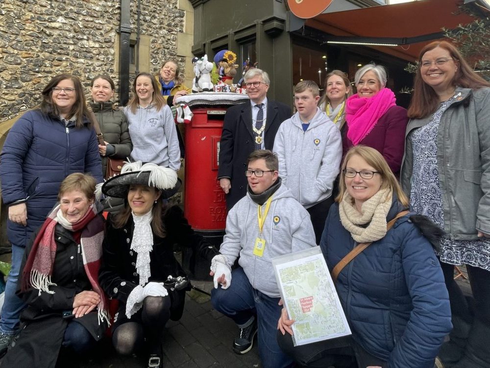 The launch of the 2025 Postbox Toppers fundraiser in St Albans with Clare at the front with her map, and the town centre Postbox at the back, in the middle. It is surrounded by the charity groups, topper makers, dignitaries and Daisy Cooper MP.